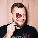 Portrait of a man smiling and holding a heart-shaped lollipop in front of a curtain backdrop.
